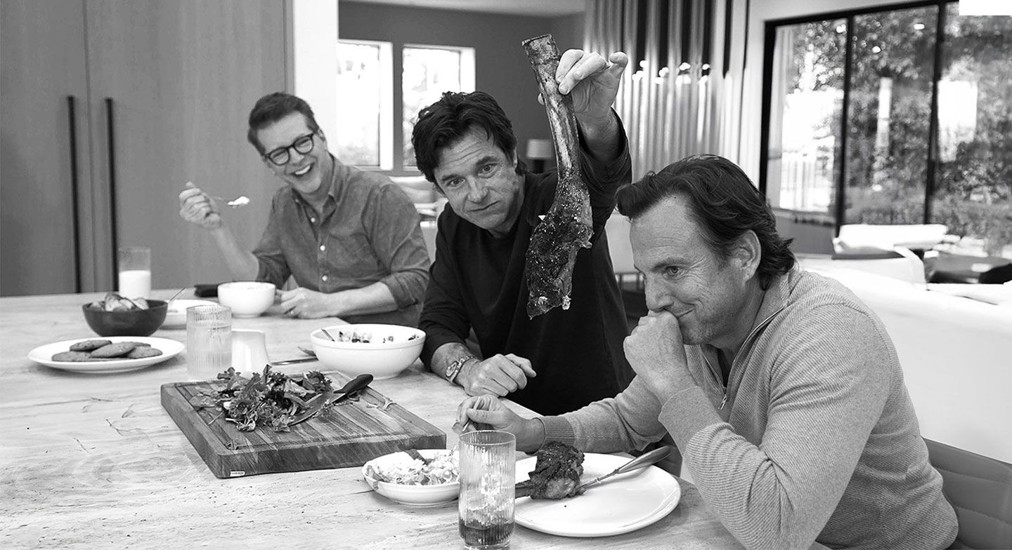 Jason Bateman holds up a giant T-bone steak while Sean Hayes laughs in the background.